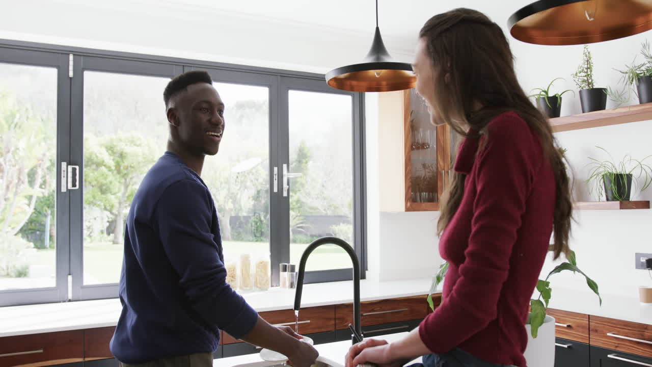 Happy diverse couple washing dishes, taking and laudhing in kitchen,slow motion