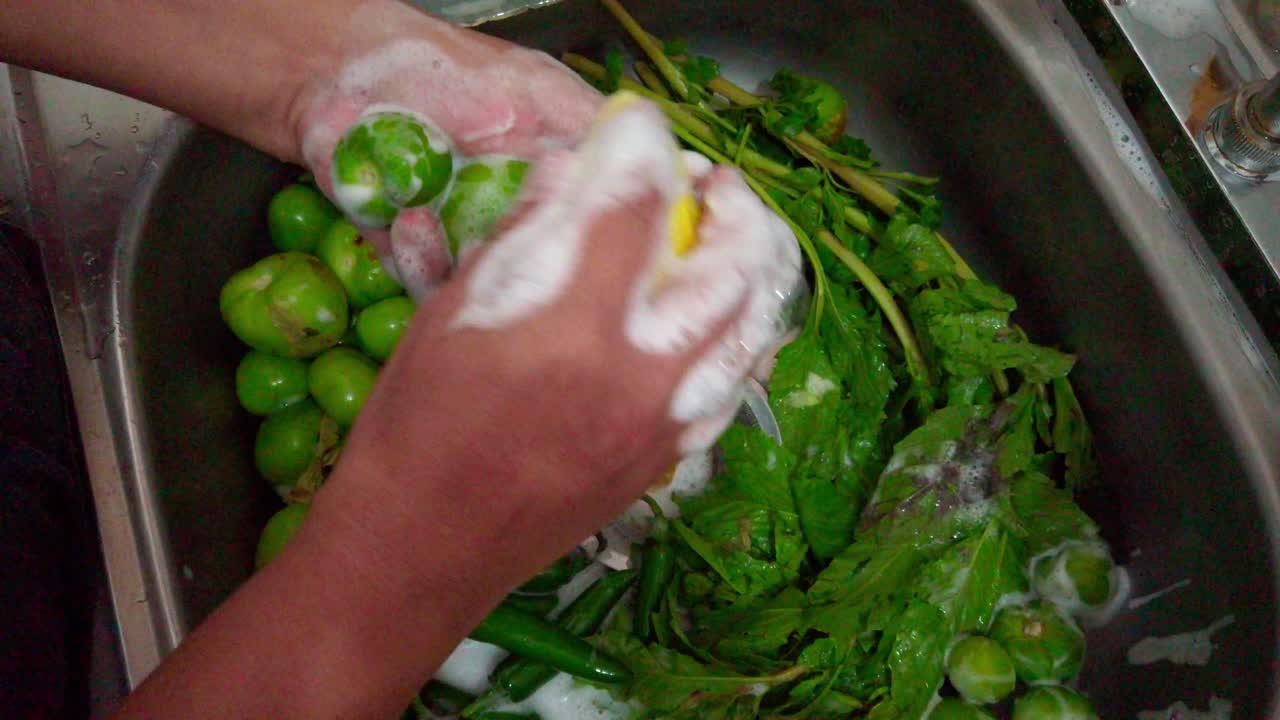 Top-down close-up of hands washing green limes and herbs with soap and sponge in a kitchen sink