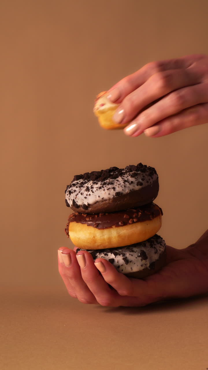 Close up of hands holding stacked donuts while top vanilla glazed donut with colorful sprinkles is lifted away. Chocolate iced and cookie crumb donuts below. Warm beige studio background