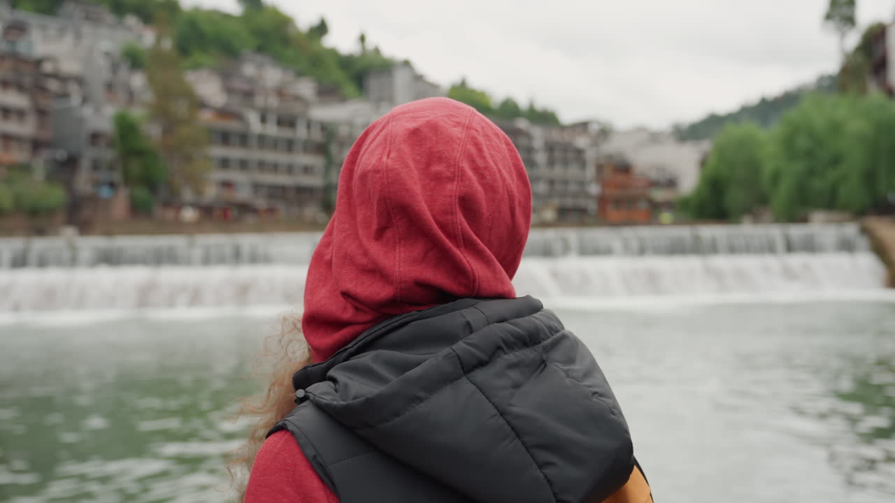 Jovem mulher asiática à beira do rio refletindo, vestindo moletom vermelho e boné amarelo com óculos e mochila em passarela de madeira, cachoeira e vila histórica ao fundo, céu nublado, pensativa e em ritmo lento.