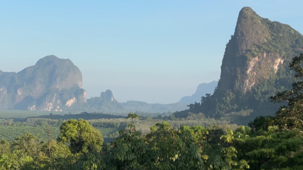 Tayland'ın Krabi bölgesindeki Din Daeng Doi'de gün doğumu sırasında yemyeşil ormanlar ve yükselen kireçtaşı dağları boyunca yapılan kaydırma çekimi; berrak gökyüzü, yumuşak ışık ve doğal tropikal manzara eşliğinde.
