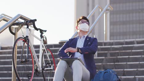 Asiatischer-Mann-Mit-Gesichtsmaske-Und-Laptop-Sitzt-Auf-Der-Treppe-Im-Firmenpark