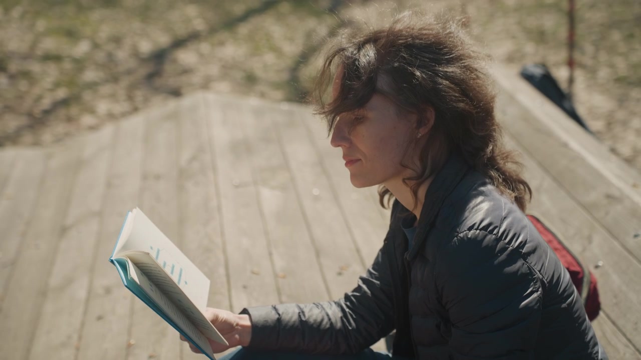 Primo piano di una donna che legge un libro all'aperto su una panchina di legno, con una borsa rossa e uno zaino nelle vicinanze. Pagine del libro visibili, luce solare che proietta ombre morbide in un ambiente naturale.