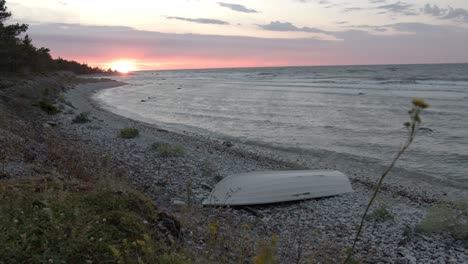 Baltic-seashore-during-sunset,-waves-hitting-the-rocky-beach-with-a-small-rowboat-in-the-bottom-of-the-frame