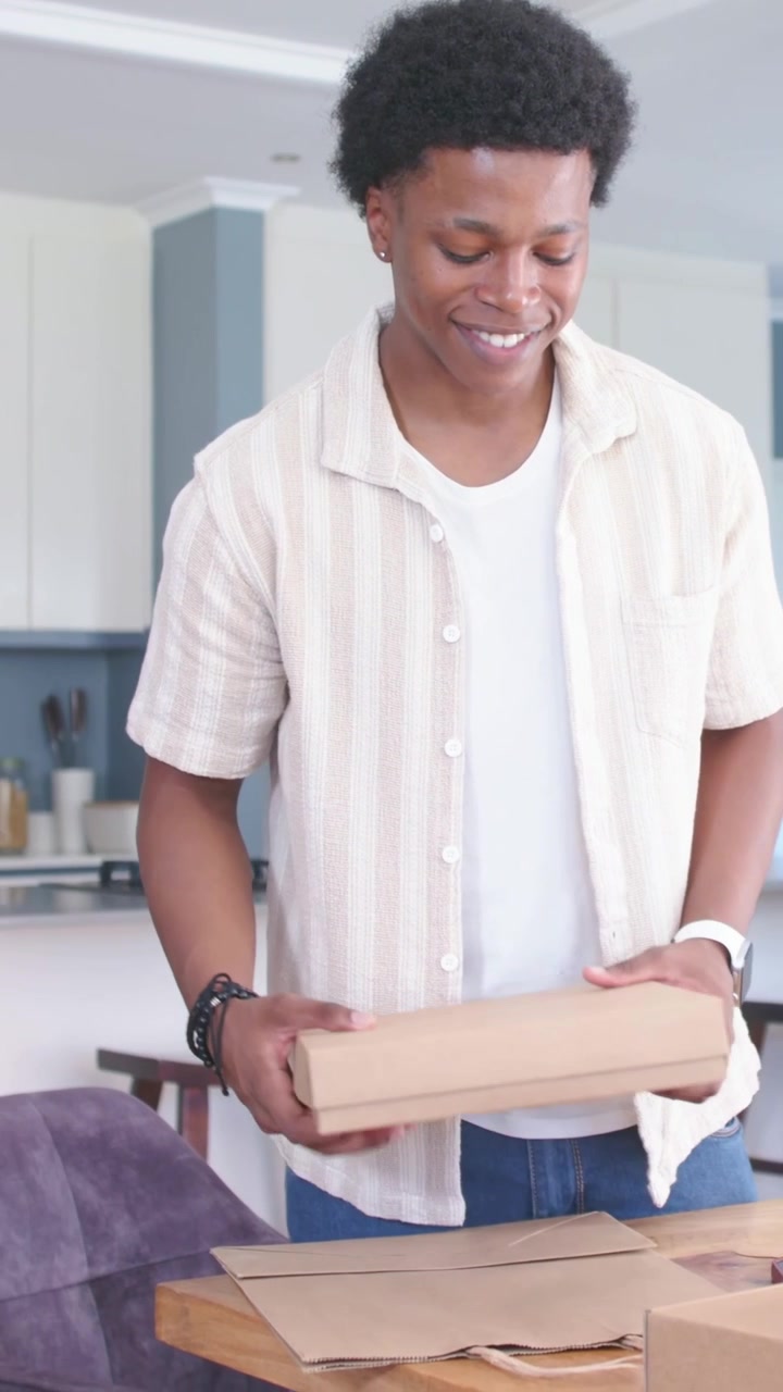 Vertical video: Smiling man in striped shirt folding boxes on wood table in kitchen, packing orders