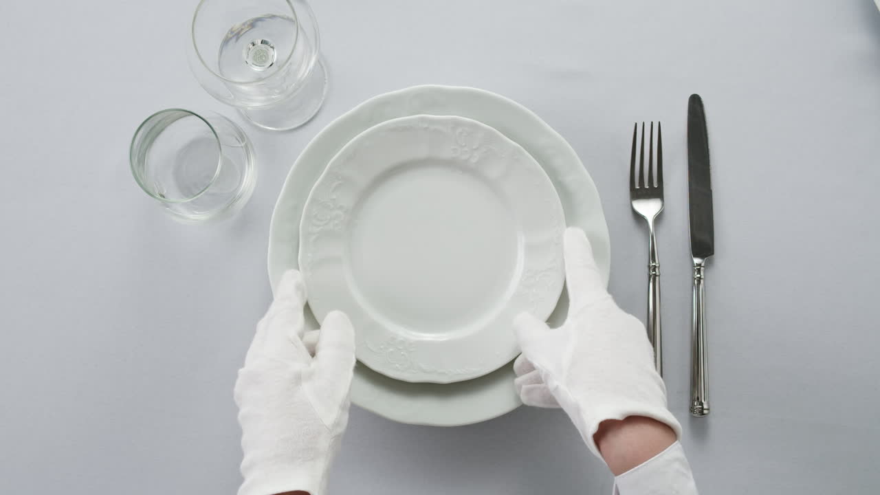 Professional table setting with white gloves. Plates, fork, knife, and glass arranged on a white table.