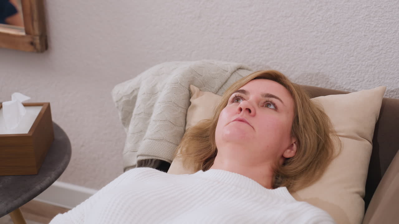 Mindful partner lies on comfortable sofa facing upward conversing with counselor, pillows support head, table nearby holds clock, tissue box and pencils, carpet, warm therapy setting