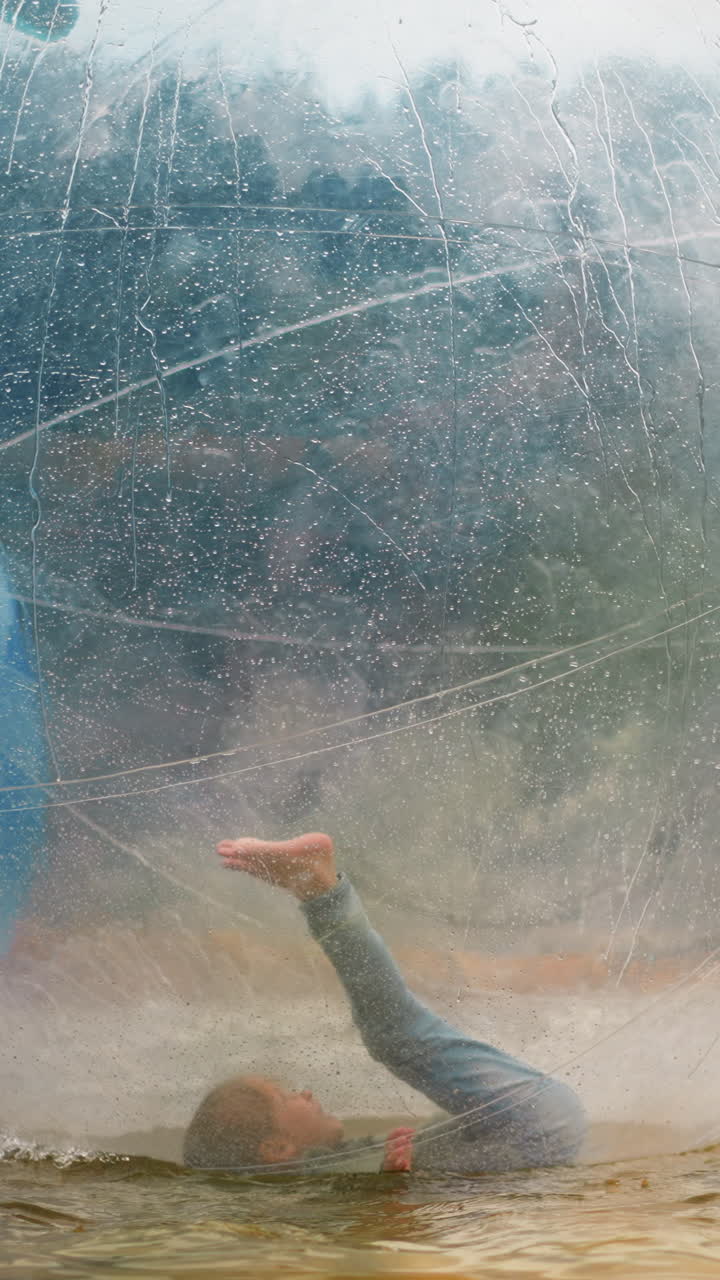 Funny little girl falls down inside large transparent water balloon floating in small pool at resort in forest on spring day slow motion