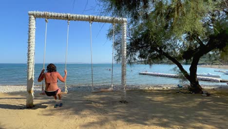 vista trasera de una mujer adulta en vacaciones de verano disfruta columpiándose en un columpio de cuerda mirando el mar en calma