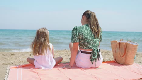 a mother and daughter sitting back to back on an orange towel in the sand by the sea on a sunny day with their hair blowing in the wind