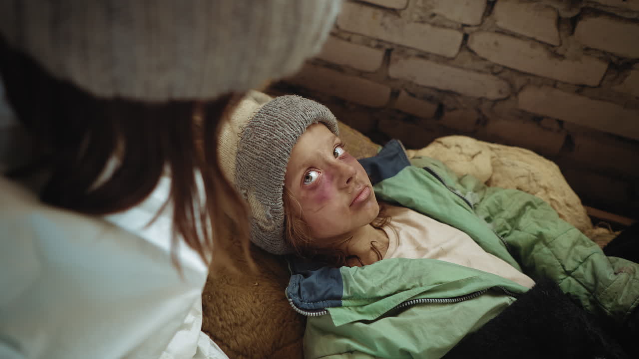 Close up of stray girl with bruised face lying on mattress inside ruined shelter, dressed in worn winter jacket and beanie, looking up at caregiver with tired eyes expressing pain in post apocalypse