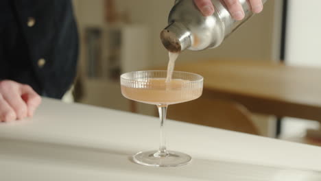Pouring-drink-from-cocktail-shaker-into-beautiful-champagne-drinking-glass-at-home-in-the-kitchen