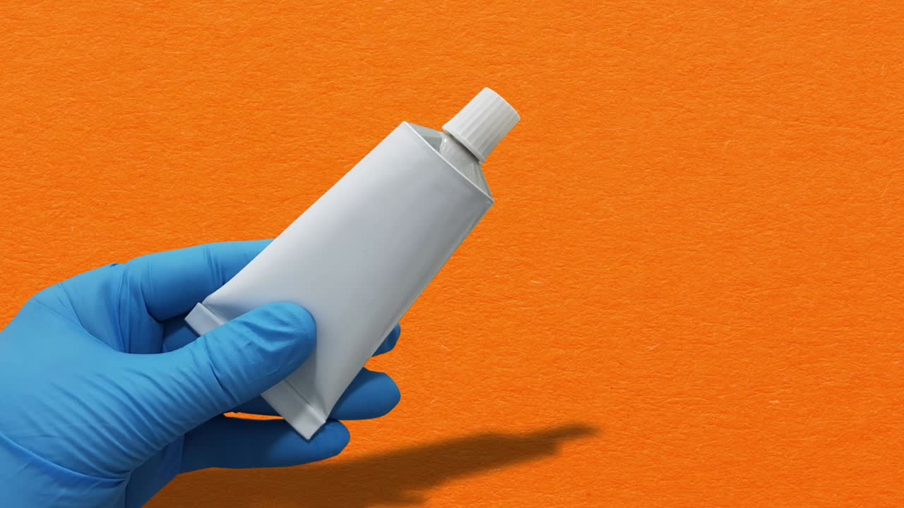 Doctor's hand wearing blue glove holding a white tube of ointment against an orange background