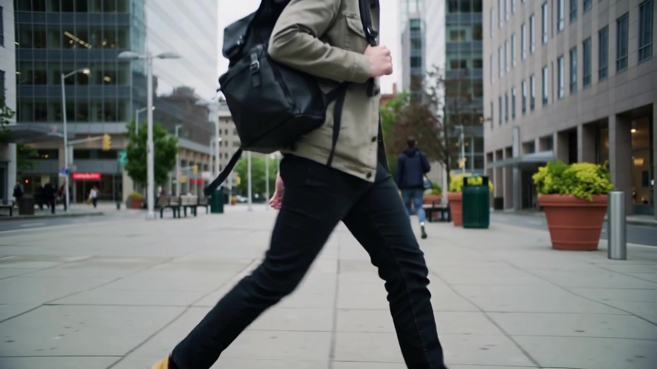 Entering plaza, adult man in light jacket holding black backpack walking to entrance passing posts