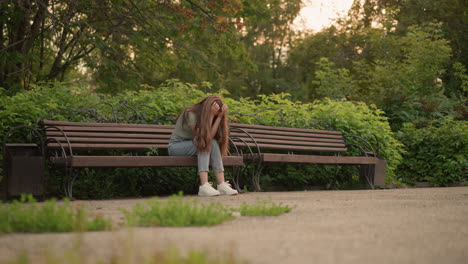 woman seated on wooden bench in reflective, somber posture with hands gently covering her face, long blonde hair cascades down her shoulder as she sits surrounded by lush greenery