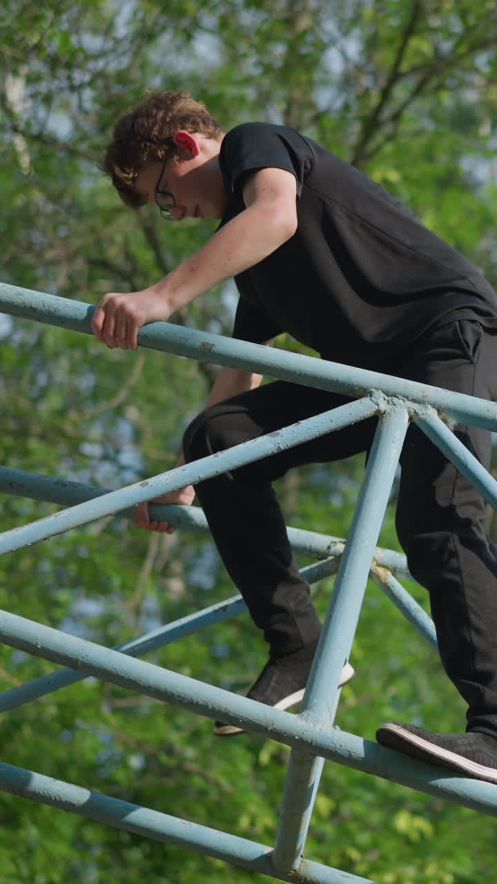 un niño joven con un traje negro y gafas se mueve cuidadosamente hacia atrás en equipos al aire libre azules, con árboles verdes exuberantes en el fondo