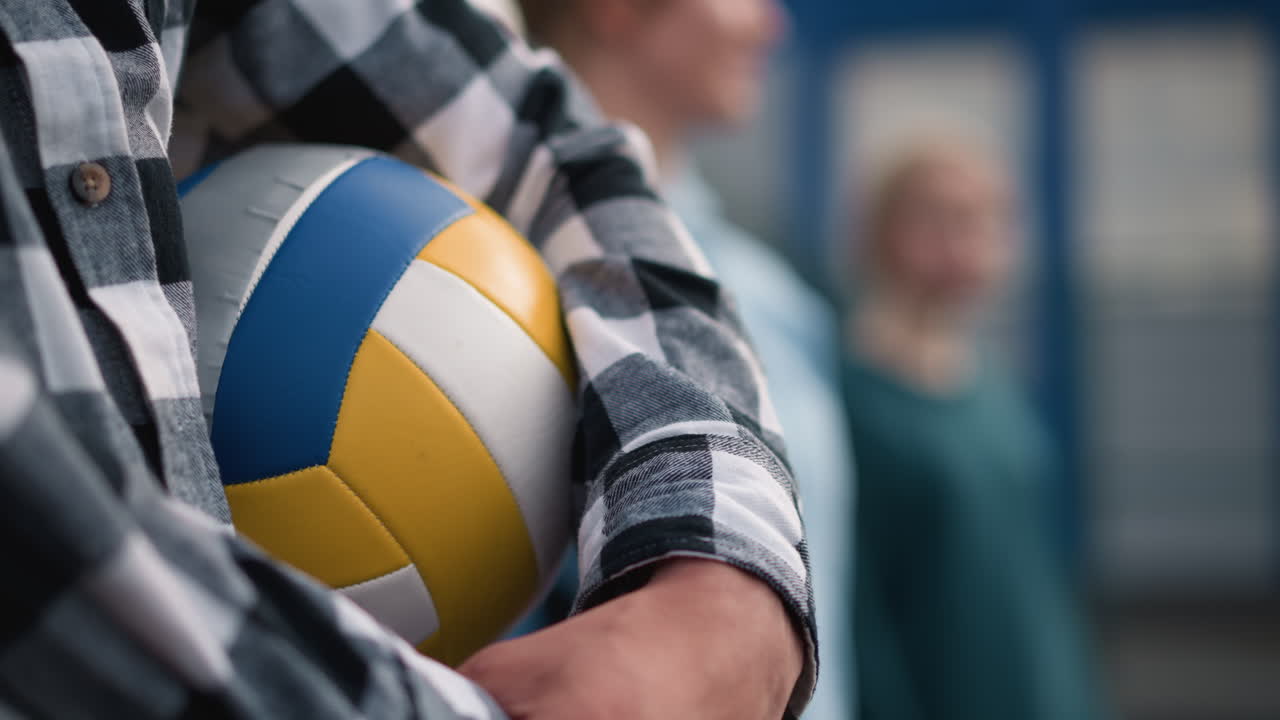 배경에는 두 명의 흐릿한 여성이 배경에 가까이 서있는 배구 공을 들고 있는 체크 셔츠를 입은 남자의 클로즈업, 배경에 부드러운 초점은 주제와 스포츠 분위기를 강조합니다.