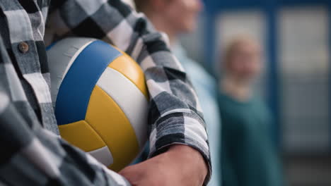 primer plano de un hombre con camisa a cuadros sosteniendo una pelota de voleibol, con dos mujeres borrosas de pie cerca de él en el fondo, el enfoque suave en el fondo hace hincapié en el tema y la atmósfera deportiva