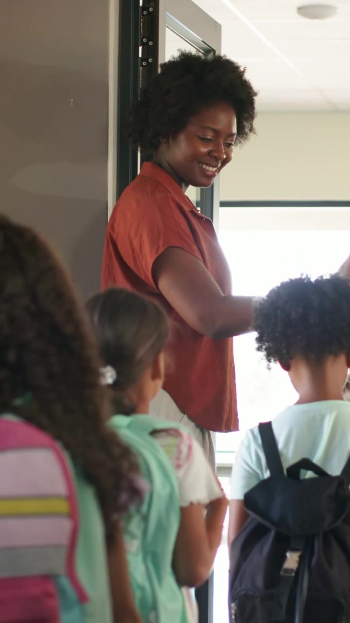 Video verticale: Un'insegnante con un top color ruggine che saluta e guida i bambini in una stanza mentre gli zaini passano attraverso l'ingresso.