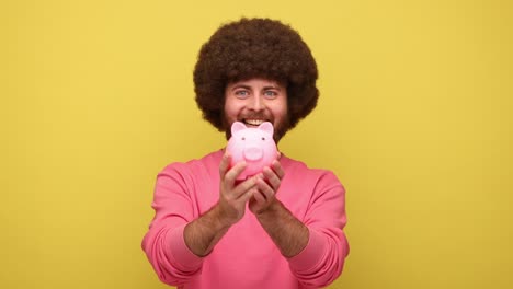 bearded hipster man holding shaking piggybank, looking at camera and showing thumb up.