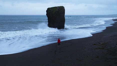 Mujer-Con-Chaqueta-Roja-Parada-En-La-Playa-De-Arena-Negra-De-Laekjavik-En-Oceanside,-Islandia