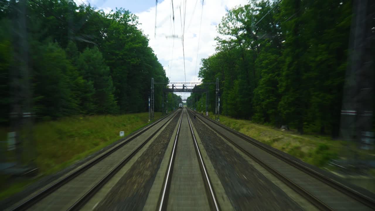 울창한 초록색 나무와 풀로 둘러싸인 기차 트랙은 공중 전선으로 둘러싸여 있습니다. 멀리 뒤에서 보이는 다른 기차가 다른 도시로 향하고 있습니다.