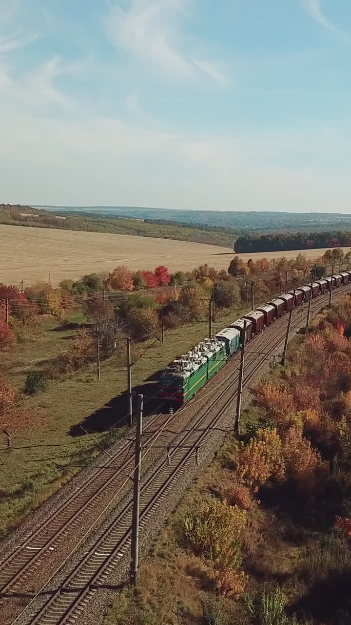 freight train is moving by rail to the nearest town on the background of the countryside with fields and road. Aerial view Vertical video