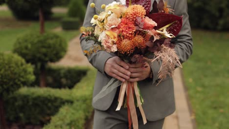 Groom-goes-down-the-alley-between-bushes-with-a-wedding-bouquet-to-his-beloved-bride,-first-meeting