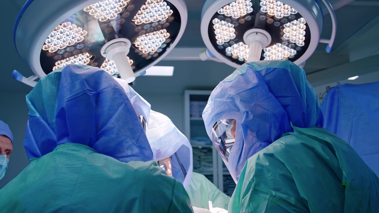 Due medici professionisti con caschi protettivi al lavoro durante un'operazione. Vista posteriore di chirurghi in piedi sotto lampade rotonde in sala operatoria.