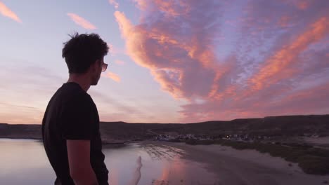 young man wearing sunglasses looking at the small seaside village next to the beach and sea at sunset