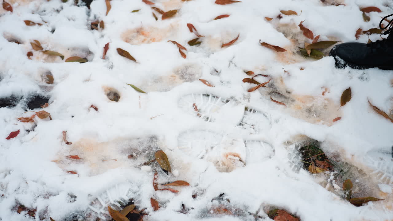 검은색 부츠를 입은 사람이 얼음으로 뒤인 얼음으로 인 땅에 활기차게 뛰어다니며 어진 건조한 잎과 여러 발자국을 가진 근접 다리 시각