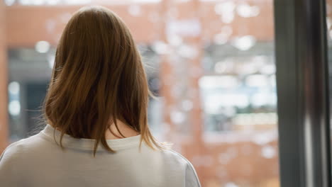 vista trasera de una mujer joven con cabello marrón hasta los hombros con una sudadera blanca, caminando en un fondo suavemente iluminado con patrones borrosos de arquitectura moderna y reflejos de luz natural