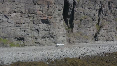 a white suv offroad vehicle drives along a rough gravel road along the base of a steep rocky cliff