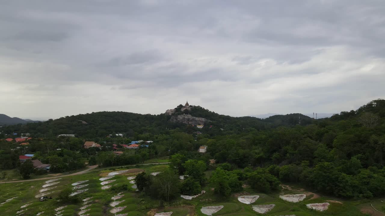 A reverse aerial footage over the edge of a forest revealing a temple on the hill and a Chinese cemetery; Wat Phra Phutthachai and a Chinese cemetery, Saraburi, Thailand