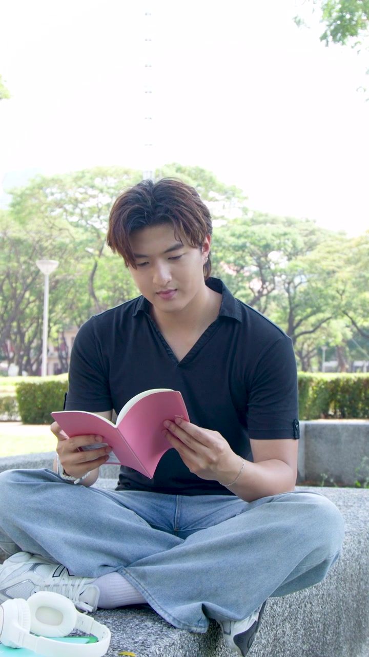 ชายหนุ่มชาวไทยนั่งขัดสมาธิกลางแจ้ง กำลังอ่านหนังสือสีชมพูภายใต้แสงธรรมชาติที่สดใส