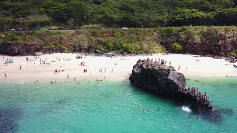 drone shot moving towards a group of people cliff jumping off of the rock at waimea bay, pupukea, hawaii