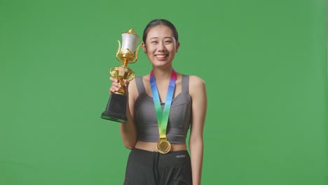 asian woman wears a gold medal and holds a gold trophy in her hand as the first winner smiling to camera on green screen background in the studio