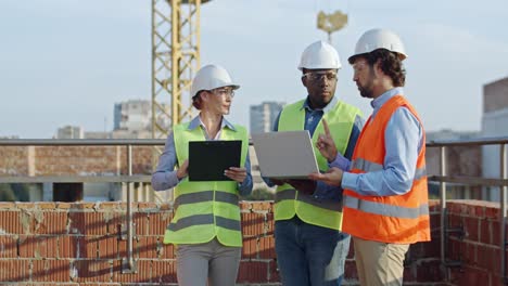 three young mixed-races builders and architects, two men and woman standing together at the constructing site with laptop computer and tablet device and discussing work process.