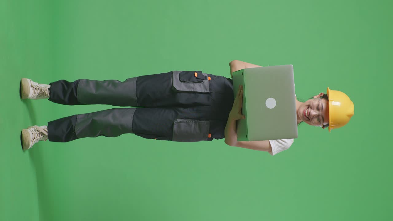 cuerpo lleno de trabajadora asiática que usa gafas de protección y casco de seguridad usando una computadora portátil mientras está de pie en el estudio de fondo de la pantalla verde