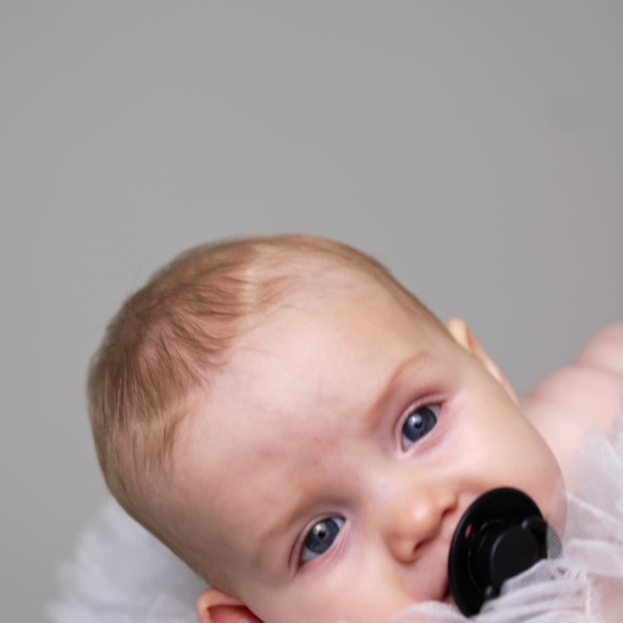 Face of an adorable grey-eyed Caucasian baby with pacifier. Infant swayed in mom's hands falling asleep. Close up