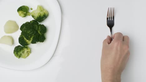 Vista-De-Arriba-Hacia-Abajo-Lugar-De-La-Mano-De-La-Persona-Y-Quitar-El-Tenedor-De-La-Mesa-Con-Brócoli-Y-Plato-De-Patata,-Mesa-Blanca