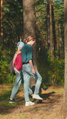 vista lateral de dos mujeres caminando a través de un denso bosque iluminado por el sol, una con corbata de cabello azul y mochila, otra con tela drapeada sobre la bolsa, el cabello atado hacia atrás, la luz del sol se filtra a través de los árboles