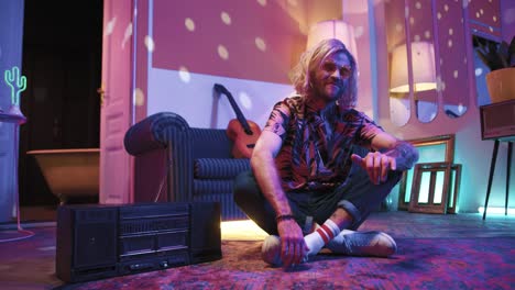 young stylish caucasian man with long hair sitting on the floor and dancing in a retro party at home