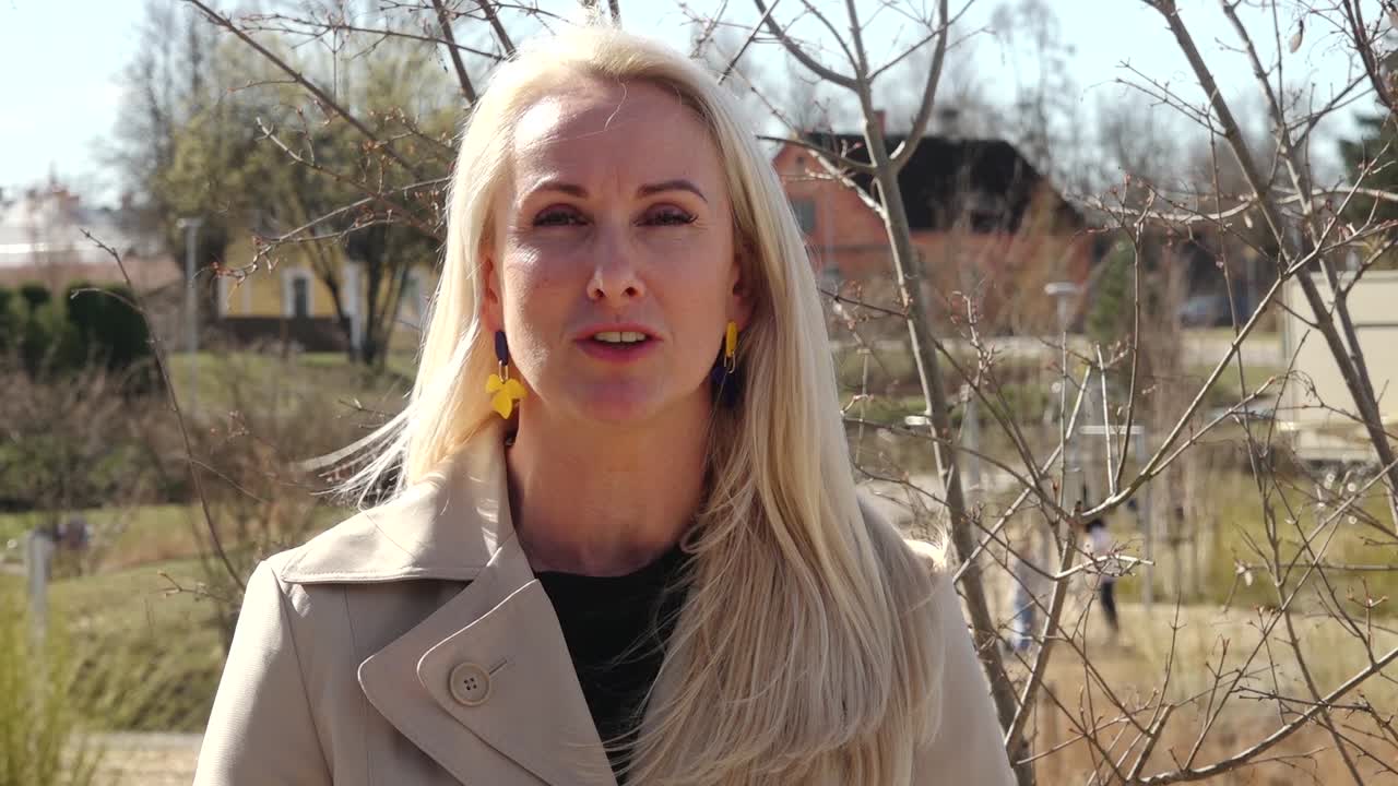 A woman with long blonde hair and colorful earrings smiles confidently while standing outdoors in a beige coat, with trees and buildings in the background.