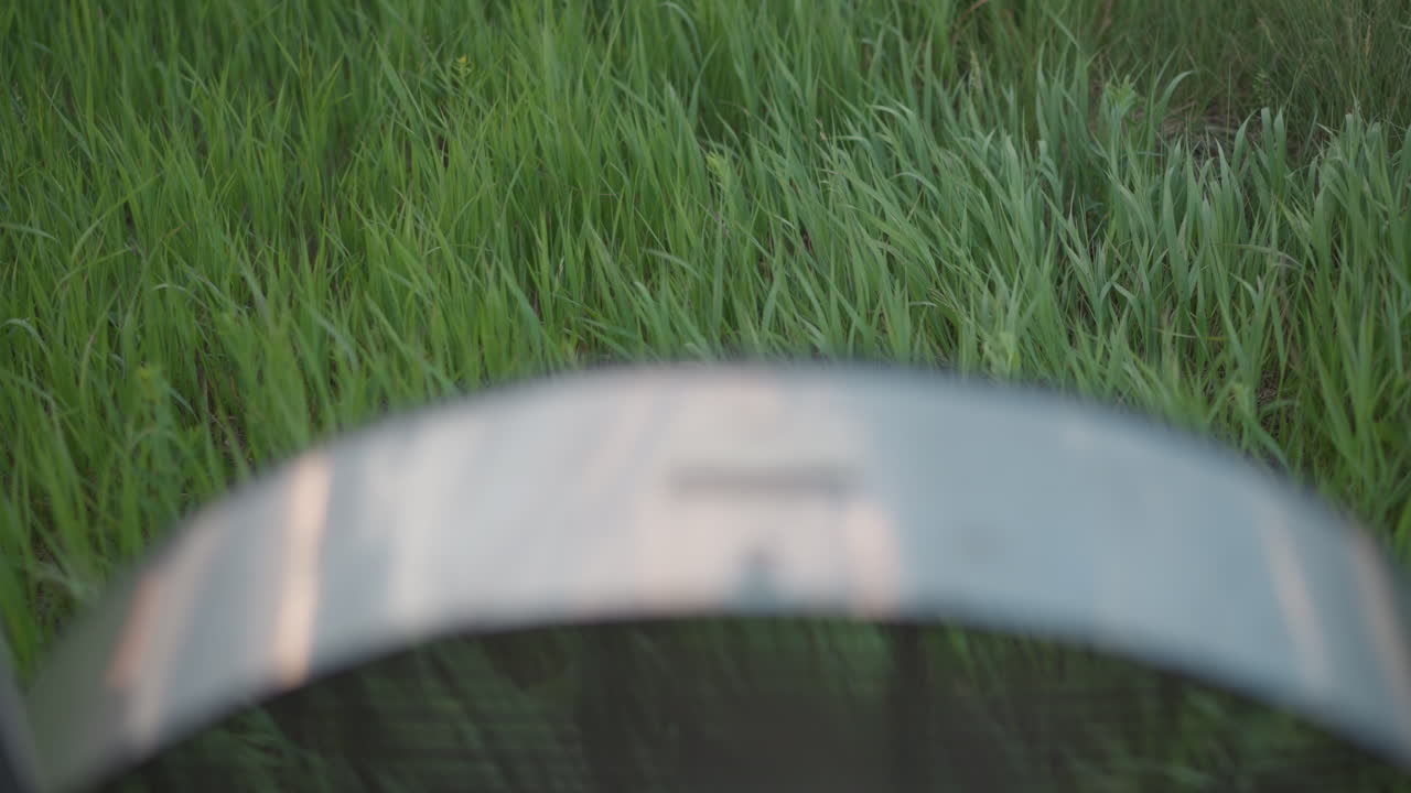 closeup slow motion view of rotating metal fan rim partially out of focus in foreground while tall green grass sways under evening breeze during outdoor equipment setup