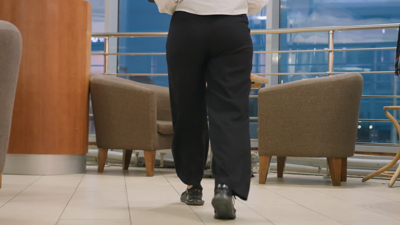 Girl walks into cafeteria and approaches seat near window, placing coffee and gadget on wooden table with glass panel behind her reflecting evening city environment and soft indoor lighting