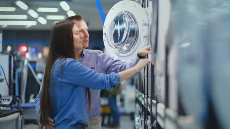 young married couple man and woman in appliances store choose to buy a washing machine for the house. open the door looking into the drum, compare the design and characteristics of the devices.