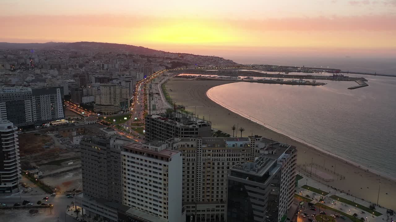 Ampia ripresa aerea che cattura la curva della Baia di Tangeri al tramonto. Presenta moderni grattacieli lungo la Corniche, la spiaggia sabbiosa e il calmo Mar Mediterraneo nel Marocco settentrionale.