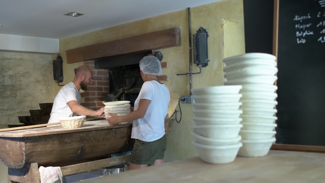 Large shot, background, a baker flouring, cuting and puting bread loaf in a traditional stone oven, using baker’s peel, foreground a women is managin bread’s baskets