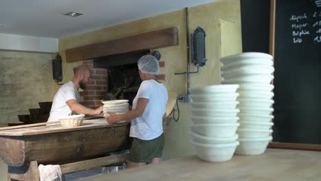 large shot, background, a baker flouring, cuting and puting bread loaf in a traditional stone oven, using baker’s peel, foreground a women is managin bread’s baskets